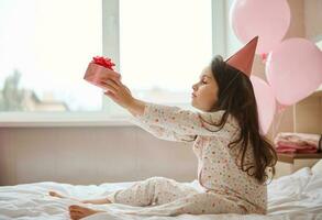 Adorable birthday girl in pink party hat, sitting on the bed in her bedroom and holding her birthday present in hands photo