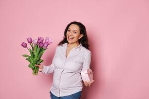 Happy smiling pregnant woman smiles, looking at camera, posing with a gift box and a bouquet of tulips, isolated on pink photo