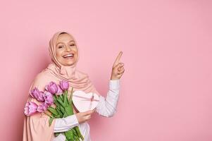 Stylish Muslim woman in pink hijab, holds a bouquet of tulips and gift box for Mother's Day, points finger at copy space photo