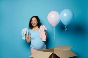 contento embarazada mujer demostración un azul y rosado recién nacido mono, aislado en azul antecedentes. género revelar fiesta concepto foto