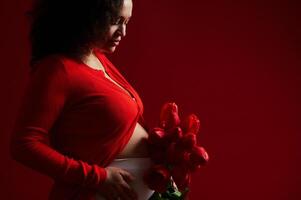 recortado ver de un embarazada mujer, expectante madre conmovedor su barriga, posando con rojo tulipanes, aislado en rojo fondo foto