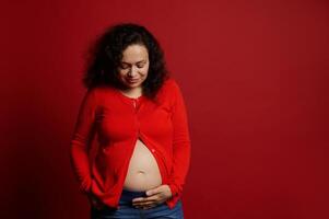 africano americano positivo embarazada mujer conmovedor su barriga, sonriente, aislado en rojo antecedentes con Copiar anuncio espacio foto