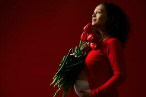 hermosa embarazada mujer en 3d semestre de el embarazo, conmovedor barriga y participación rojo tulipanes en rojo aislado antecedentes foto