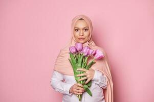 hermosa musulmán embarazada mujer vestido en rosado hiyab, participación un ramo de flores de púrpura tulipanes, con confianza mirando a cámara foto