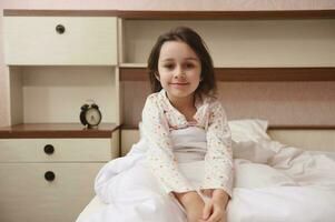 encantador pequeño niño niña en pijama, sonriente mirando a cámara, sentado en cama a su acogedor dormitorio. contento niños foto