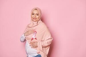 positivo musulmán embarazada mujer en rosado hiyab, participación manos alrededor un rosado satín cinta, emblema de luchando pecho cáncer foto