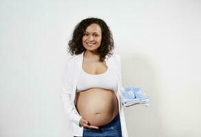 atractivo embarazada mujer participación recién nacido ropa, suavemente acariciando su barriga, sonriente a cámara. el embarazo 30 semanas foto