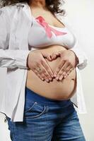 Closeup of pregnant woman wearing pink ribbon, puts hands on her belly in heart shape. Breast Cancer Awareness campaign photo