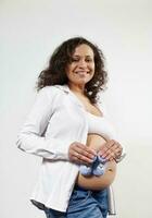 Beautiful multi ethnic pregnant woman holding blue knitted baby booties on her naked belly, isolated on white background photo