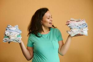 Beautiful smiling pregnant woman holding clean ironed baby clothes on her hands palms up, isolated on orange background photo