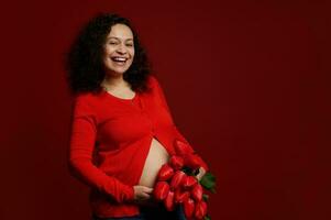 auténtico embarazada mujer con grande barriga, expectante madre, participación un ramo de flores de rojo tulipanes, aislado en rojo antecedentes foto