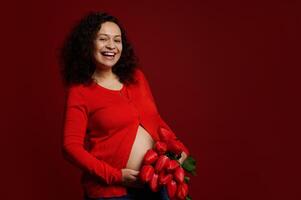 auténtico embarazada mujer con grande barriga, expectante madre, participación un ramo de flores de rojo tulipanes, aislado en rojo antecedentes foto