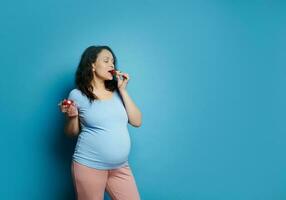 Beautiful pregnant young woman eating freah sweet strawberries out of bowl, blue background. Pregnancy. Healthy eating photo