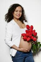 Multi ethnic pregnant woman posing bare belly with a bouquet of red tulips, smiling at camera, isolated white background photo