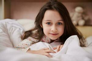 de cerca retrato de un encantador pequeño niño chica, mirando a cámara, acostado en cama con cómodo suave blanco cobija foto