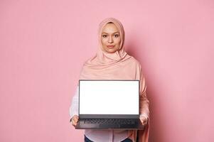 Confident Arab Muslim woman with head covered in hijab, showing at camera a laptop with white blank screen. Copy space photo