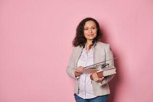 Charming positive middle aged female educator holding eyeglasses, studying books and pointer, smiles looking at camera photo