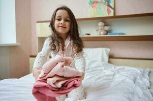 adorable pequeño niña participación fuera su doblada ropa a cámara mientras sentado en cama en su dormitorio en el temprano Mañana foto