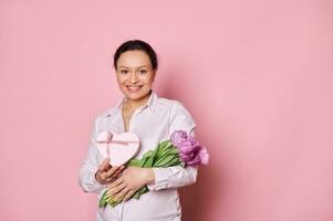 Authentic portrait on pink background of middle-aged smiling beautiful woman with gift box and bouquet of purple tulips photo