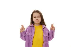 bonito pequeño niño niña con posando con cruzado dedos, esperanzas para bueno suerte y su Sueños ven verdadero, blanco antecedentes. foto