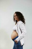 Vertical portrait of curly haired pregnant woman, posing bare belly on isolated white backdrop. Happy pregnancy 6 month photo