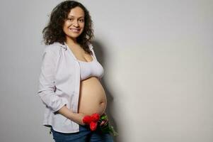 contento embarazada mujer, futuro mamá participación tulipanes en su desnudo barriga, sonriente mirando a cámara, aislado blanco antecedentes foto