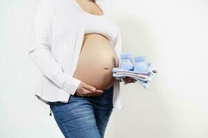 Closeup pregnant woman holding newborn baby clothes and gently caressing her bare belly, isolated over white background photo