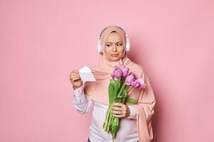 Amazed Arab Muslim woman with head covered in pink hijab, holding a bouquet of purple tulips and reading a greeting card photo