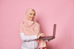 Gorgeous pregnant Arab Muslim woman in pink hijab, working online on laptop, smiling at camera, isolated pink background photo