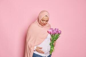 Delightful Muslim pregnant woman in pink hijab, touching tummy, expressing positive emotions, feeling first baby kicks photo