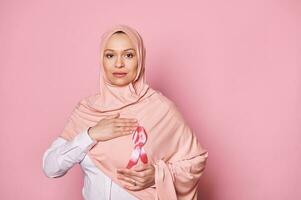 musulmán mujer en hiyab, vistiendo rosado satín conciencia cinta. internacional día luchando en contra pecho cáncer Primero octubre foto