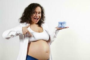 Excited pregnant pretty woman holding baby socks on her hand, pointing at her belly, rejoicing at her pregnancy 30 week photo