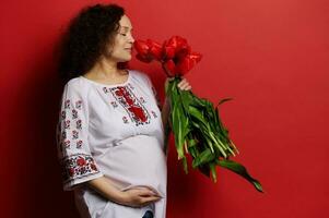 Charming ethnic pregnant woman in embroidered dress smelling bunch of tulips, gently caressing her belly, red background photo