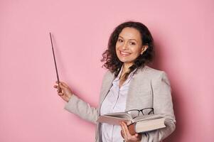 Cheerful female teacher pointing with pointer at pink background while explaining lesson, smiling looking at camera photo