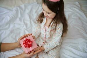 Top view amazed birthday girl in pajamas, sitting on the bed and rejoicing at a happy present from her mom for birthday photo