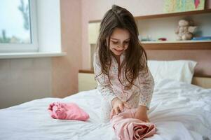 adorable pequeño niña en blanco pijama con vistoso puntos, sentado en el cama en su dormitorio y plegable su ropa foto