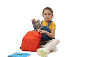 Adorable Caucasian 5 years first grader child girl puts felt-tip pens into her orange backpack, smiles looking at camera photo