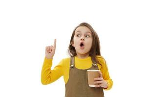 asombrado pequeño niño niña señalando su dedo a Copiar espacio en blanco fondo, posando con desechable taza de caliente bebida foto