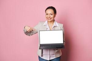 Working pregnant woman smiles at camera, points finger at white laptop blank digital screen Mockup. Pregnancy and career photo