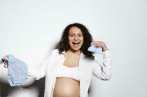 Emotional pregnant woman, holding baby clothes, smiles looking at camera, posing bare belly on isolated white background photo