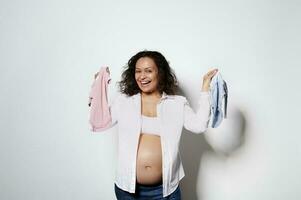 Happy pregnant woman with curly hair, holds pink and blue newborn clothes, expecting twins boy and girl, white backdrop photo