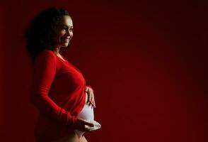 encantador adulto embarazada mujer participación blanco orquídea flor, sonriente, acariciando su barriga, expresando positivo emociones foto