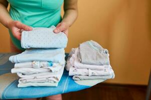 Closeup of expectant mother, pregnant woman standing by ironing board, arranging clean ironed clothes for newborn baby photo