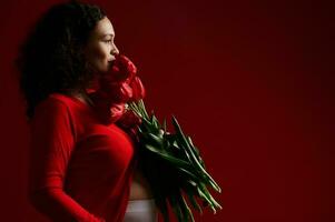 Side portrait of beautiful ethnic pregnant woman holding a bouquet with red tulips, isolated over red color background photo