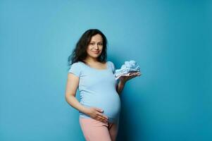 Beautiful pregnant woman holding folded ironed newborn baby laundry, touching her belly, isolated on blue background photo