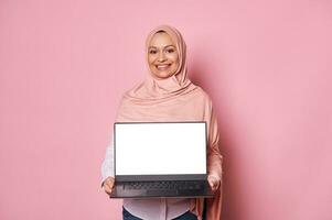 Pleasant Muslim business woman in pink hijab, smiling looking at camera, showing a laptop with white empty screen Mockup photo