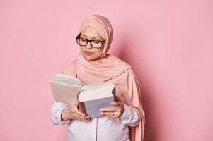 Beautiful Middle Eastern Muslim woman in pink hijab, reading book isolated on color background. Educated people concept photo