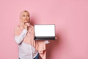 Pensive working pregnant Muslim woman in pink hijab, holds laptop with white screen, reasoning over her maternity leave photo