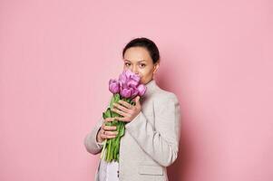 Attractive pregnant woman sniffing a beautiful bouquet of purple tulips, isolated over pink background. Mother's Day photo