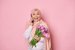 Amazed Muslim pregnant woman in hijab, holding bouquet of purple tulips, talking on mobile phone, isolated pink backdrop photo